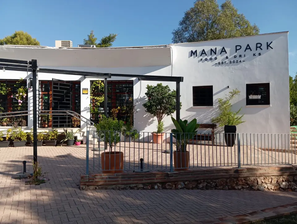 Weiß gekalkte Fassade von Mana Park mit schwarzem Schriftzug und Terrasse mit Kübelpflanzen